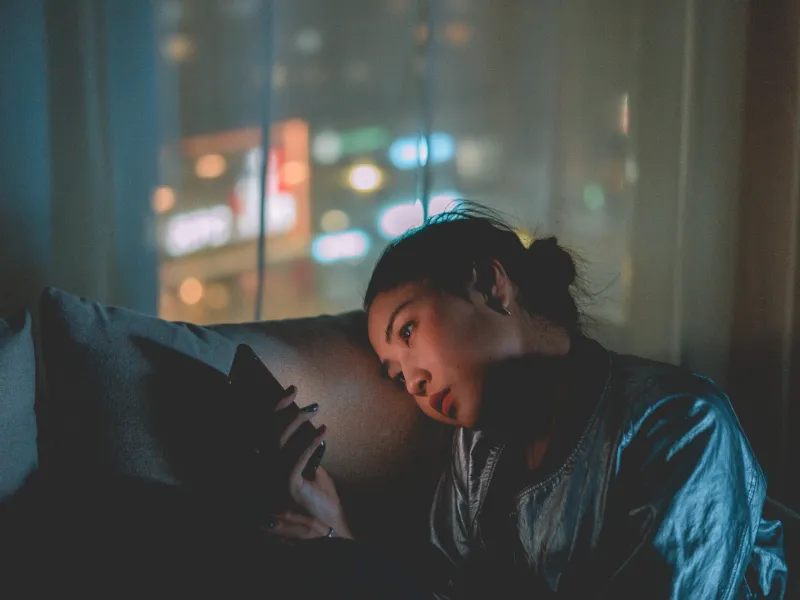 Woman using smartphone while relaxing on couch at night, city lights blurred in background