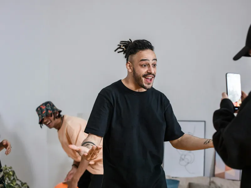 A man with dreadlocks smiles broadly as he gestures, while three friends look on; one is holding a smartphone and filming him.